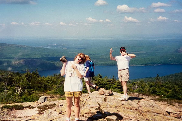 Picture of woman holding rock with older man holding arms up while standing on a rocky hill overlooking a body of water and trees in Acadia National Park